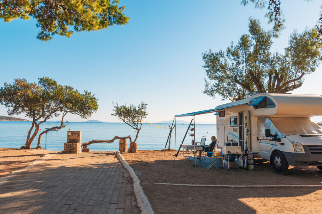 Person sitting outside an RV with awning extended at a waterfront campsite on a sunny summer day