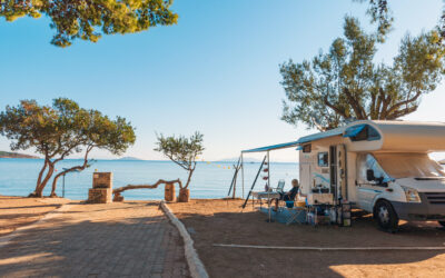 Person sitting outside an RV with awning extended at a waterfront campsite on a sunny summer day